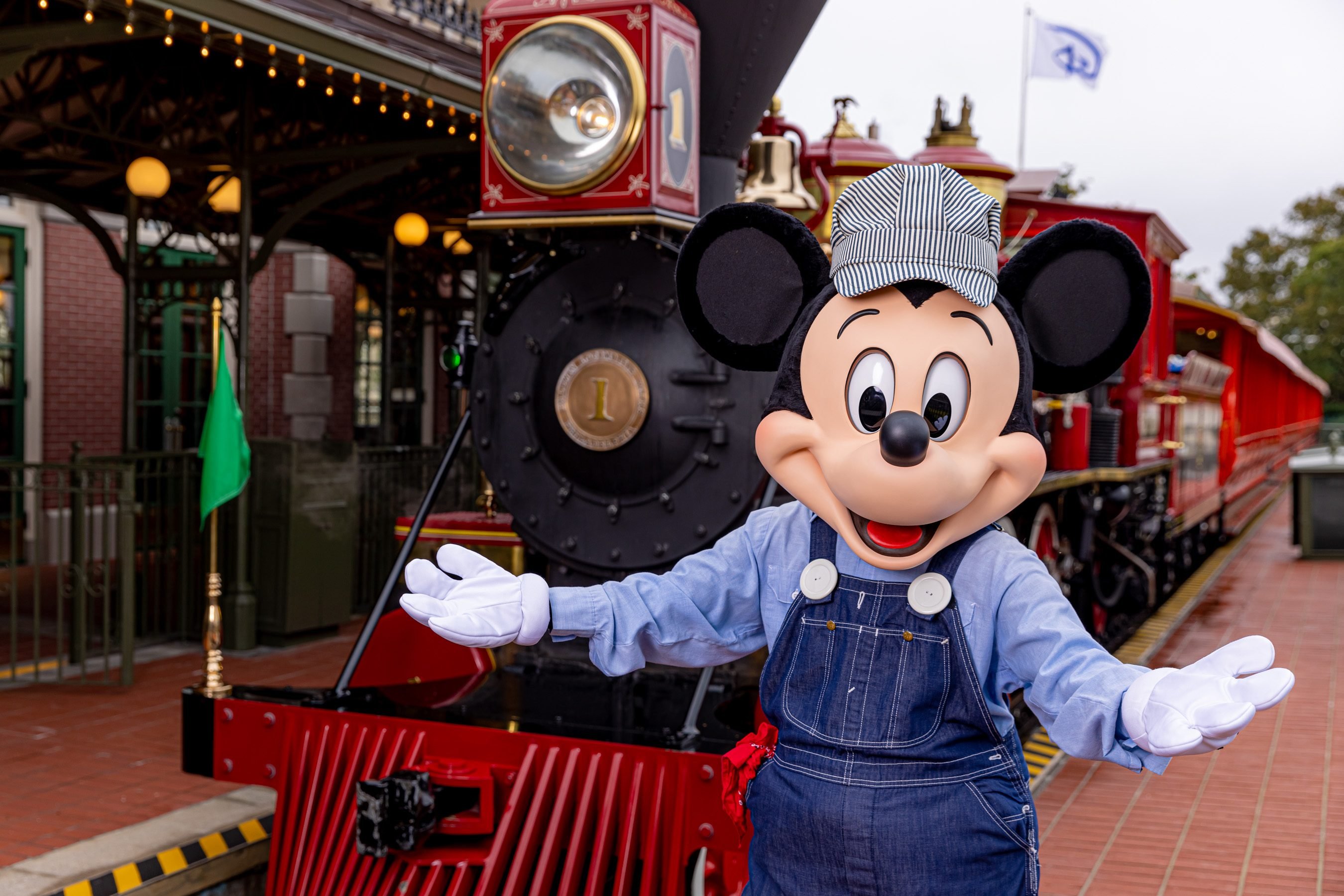 Mickey Mouse in a conductor outfit in front of a Disney theme park railroad train.