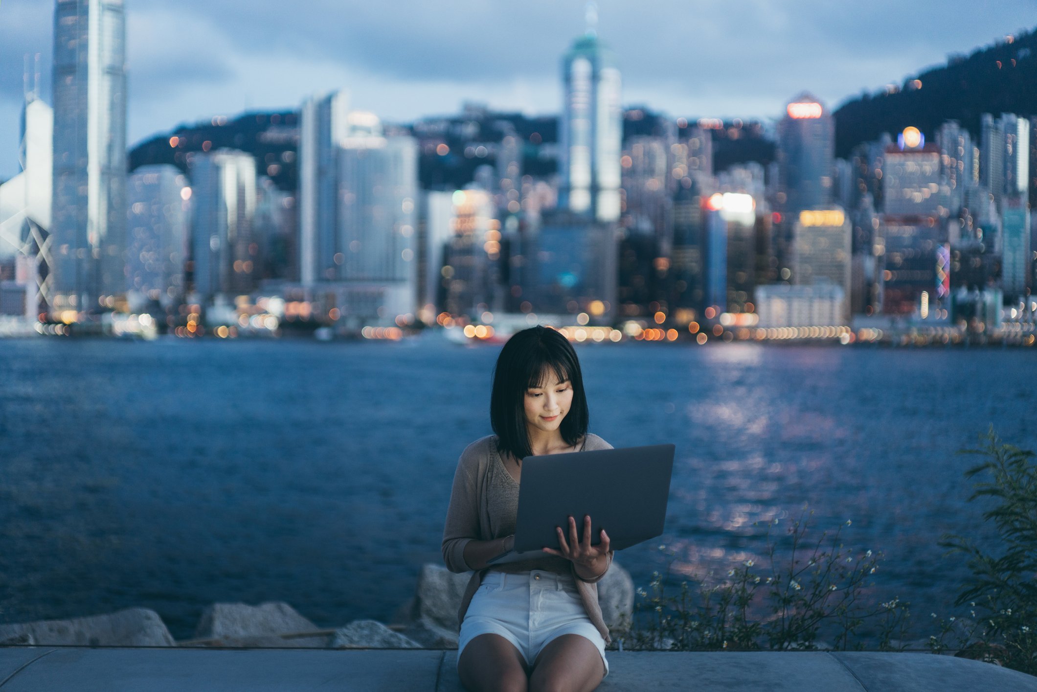 A woman looking at a laptop with the Hong Kong skyline in the background.