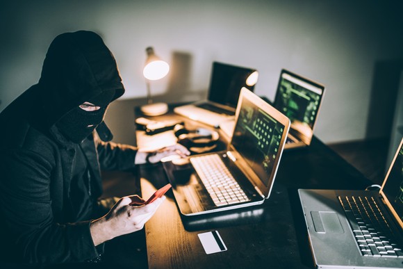 Person in ski mask looking at multiple laptop computers, mobile phones, and credit cards.