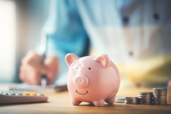 A piggy bank sitting on a desk, beside which are ascending stacks of coins.
