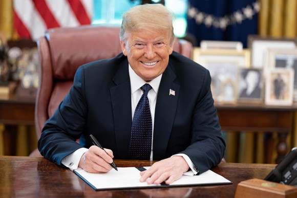 A smiling Donald Trump signing a bill while seated at a desk in the Oval Office.