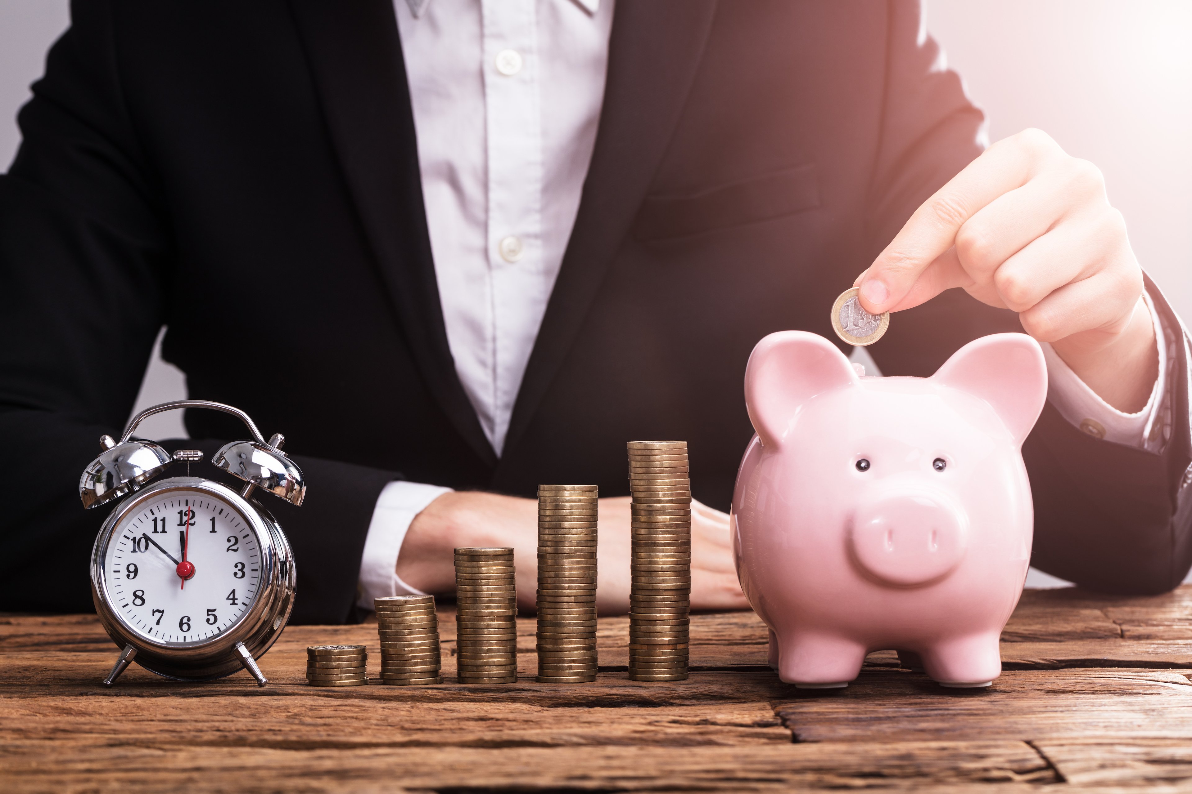 A person dropping a coin into a piggy bank, beside which are ascending stacks of coins and a clock.