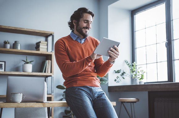 An investor leans against a desk and studies something on a tablet. 