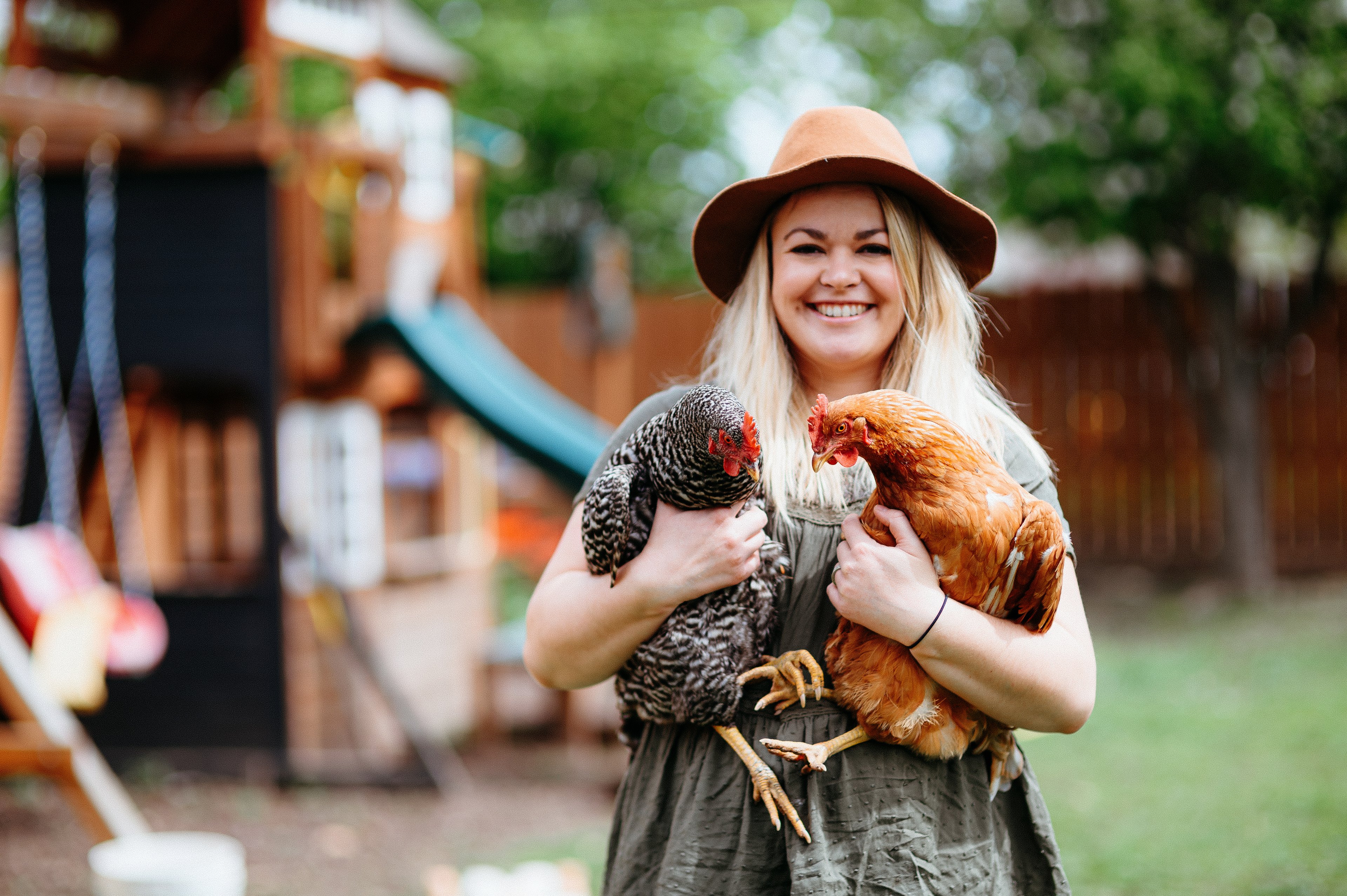Someone in a hat is smiling and holding two chickens.