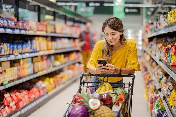 A person smiling while looking at their phone and leaning on a shopping cart full of food items in a snack food aisle in a store.