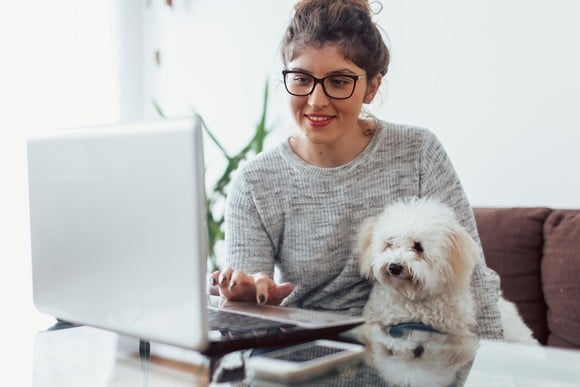 A person smiles while holding a dog in their lap and typing on a laptop computer. 