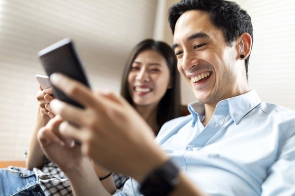 A couple is seated and smiling broadly while holding their phones.