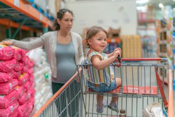 A child smiling in a shopping cart with an adult in the background at a wholesale store. 