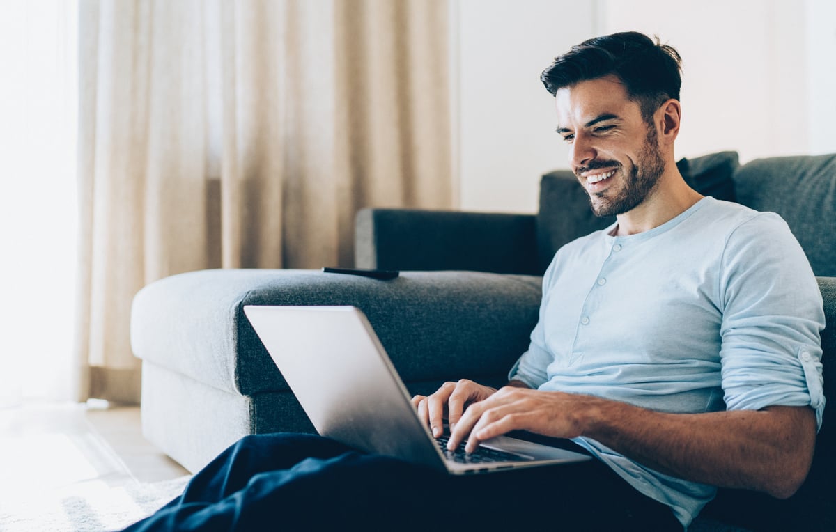 A person smiles while typing on a laptop computer and sitting against a couch. 