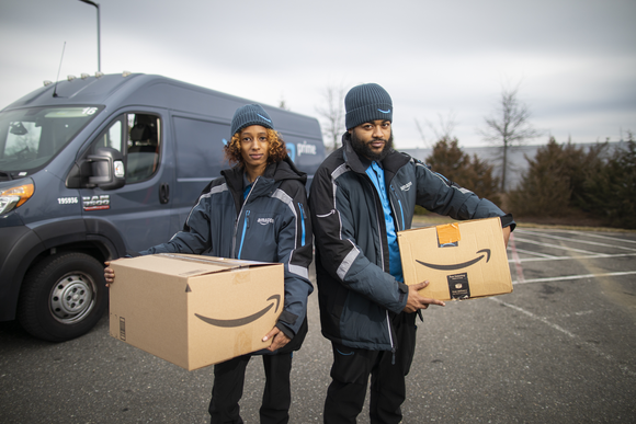 Two Amazon delivery drivers carrying cardboard boxes with the company's logo.