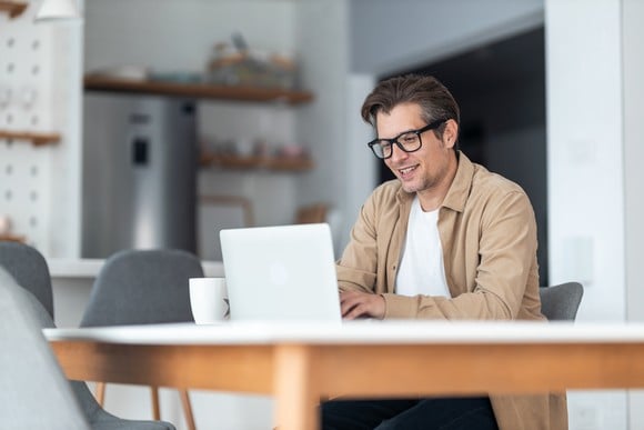 A person smiling while sitting at a table with a laptop computer and a mug. 