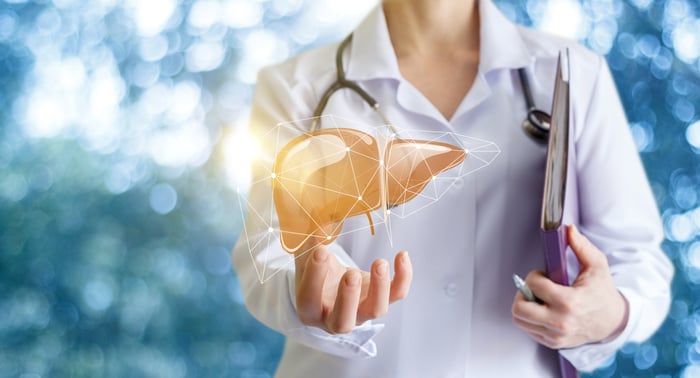 A doctor stands against a white and blue background while holding a hologram of a liver.