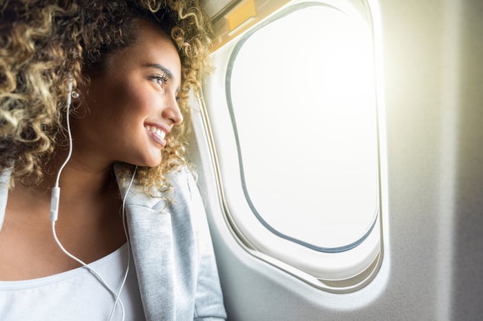 A person smiling while looking out the window of an airplane with headphones in their ears.