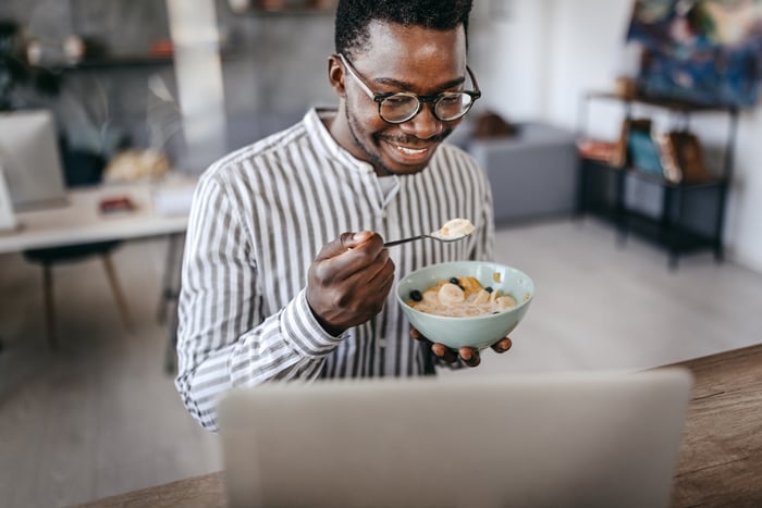 A person smiles while eating a bowl of cereal with fruit and sitting at a table in-front of a laptop computer. 