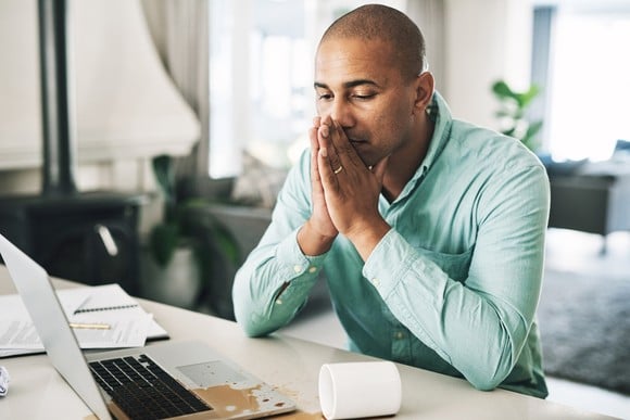 A person clasps their hands while looking at spilled coffee over a laptop computer. 