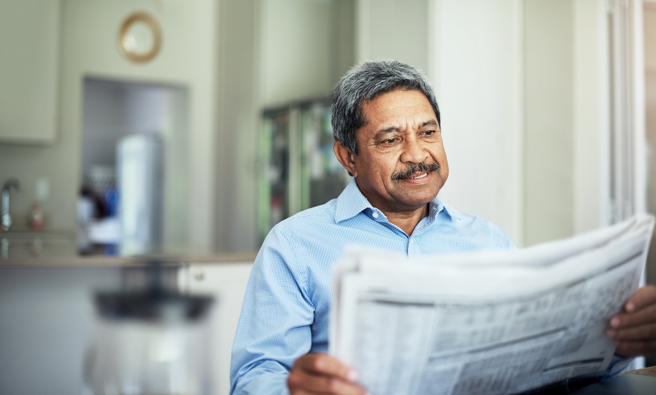 A smiling person reading a financial newspaper while seated at the kitchen table.