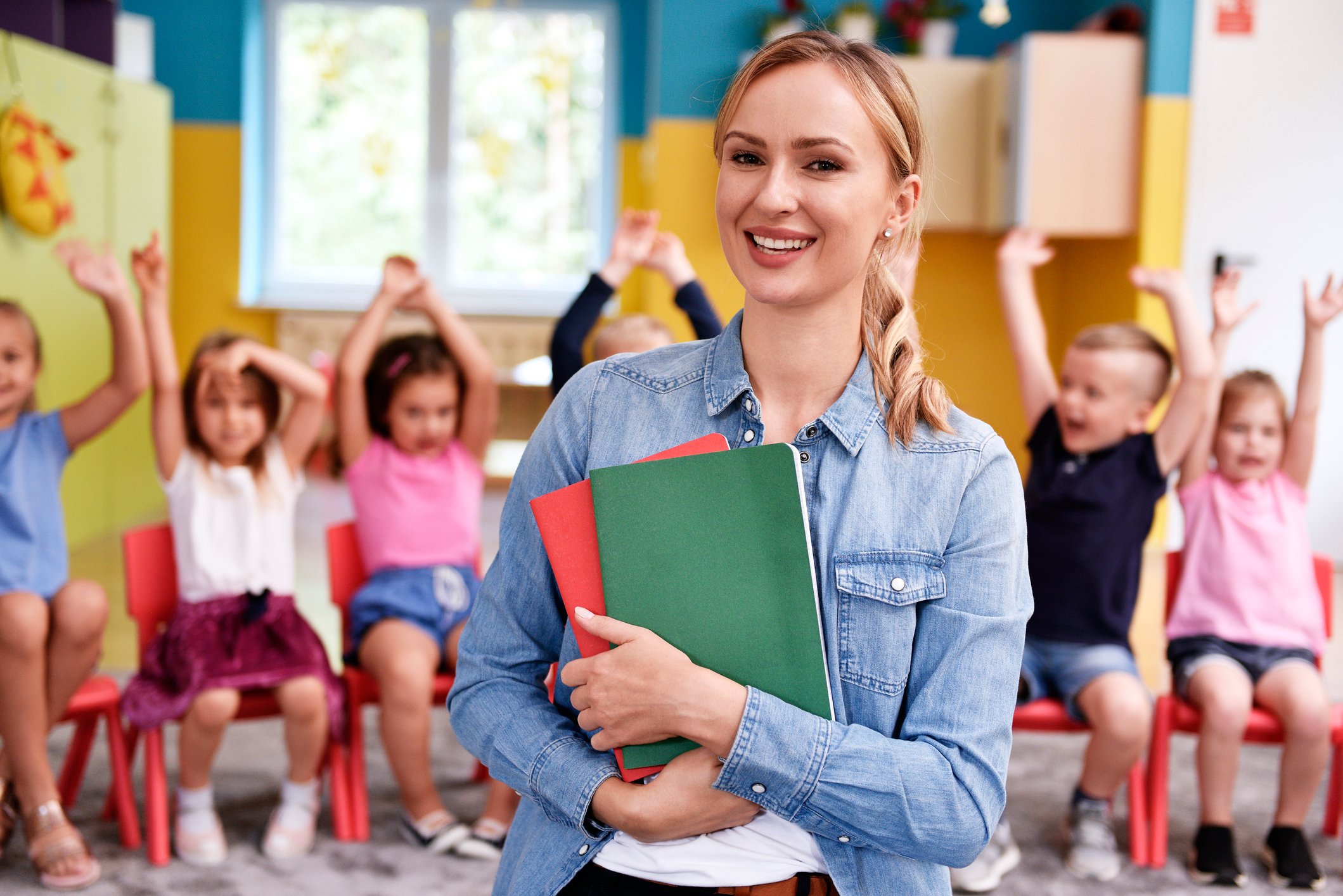 A schoolteacher is smiling with happy elementary school children behind her.