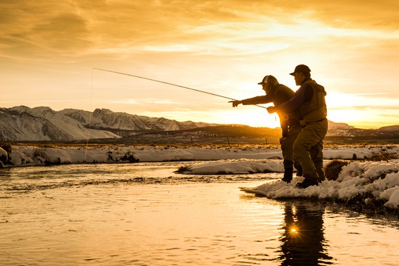 Two people fish in a stream against a yellow and orange sunny backdrop.