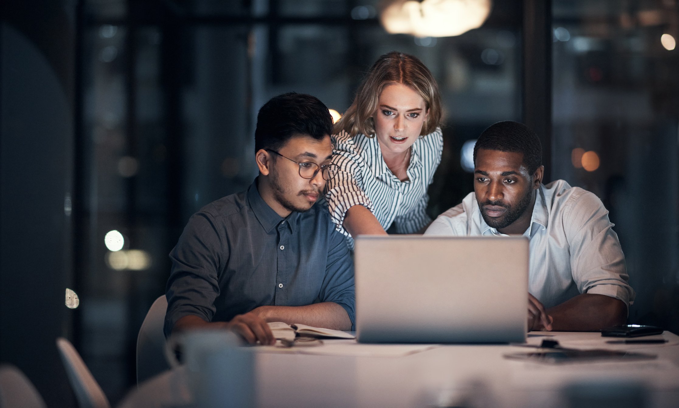 Three people look at something on a laptop in a darkened office.