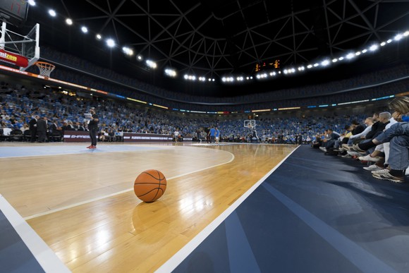 A basketball is pictured on the corner of a basketball court in a large arena.