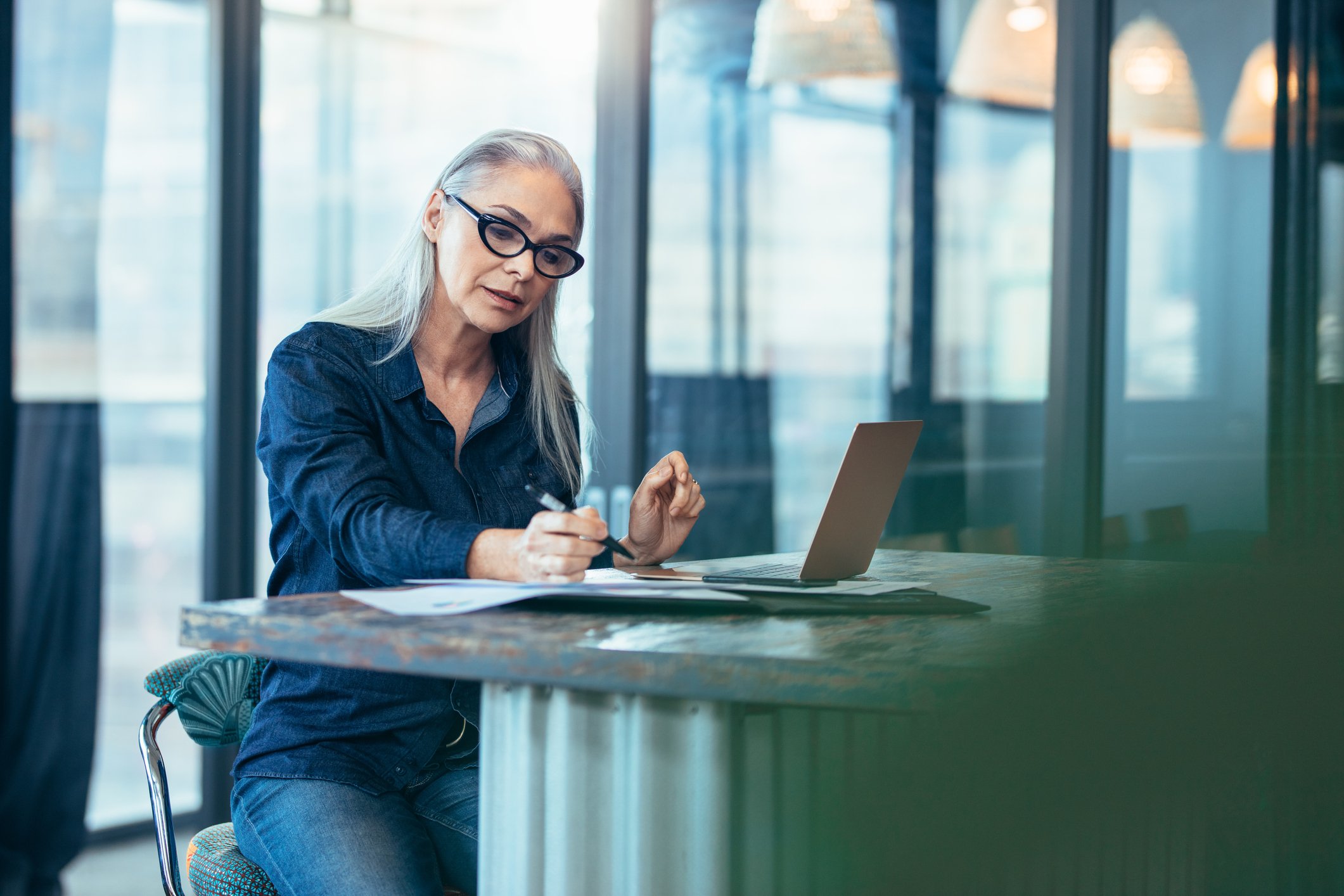 A senior woman sitting at a desk looking at papers with a laptop open.