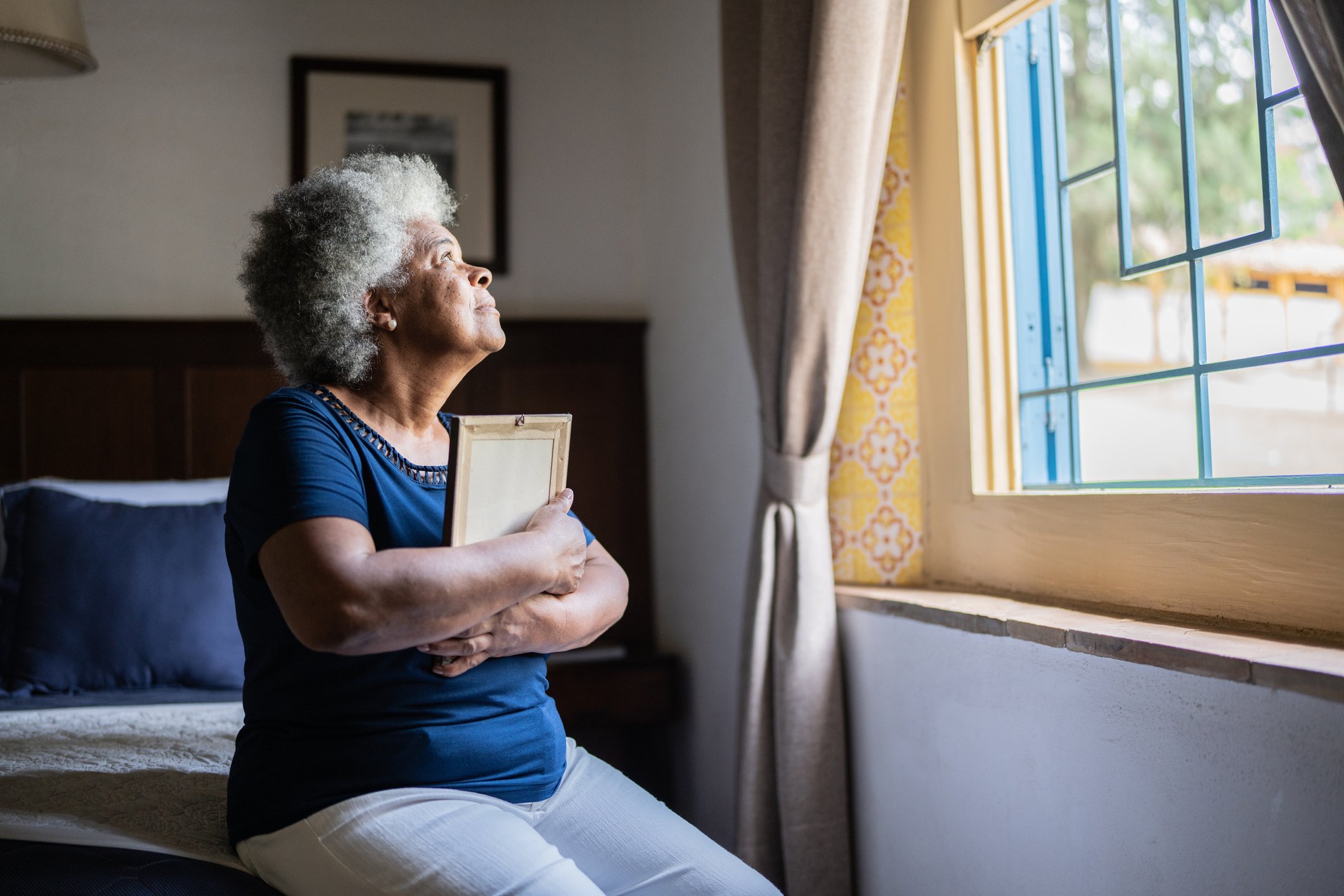Person sitting on bed, holding picture frame, and looking out window.