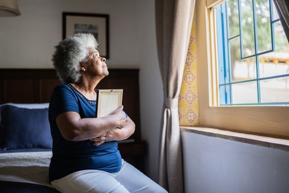 Person sitting on bed, holding picture frame, and looking out window.