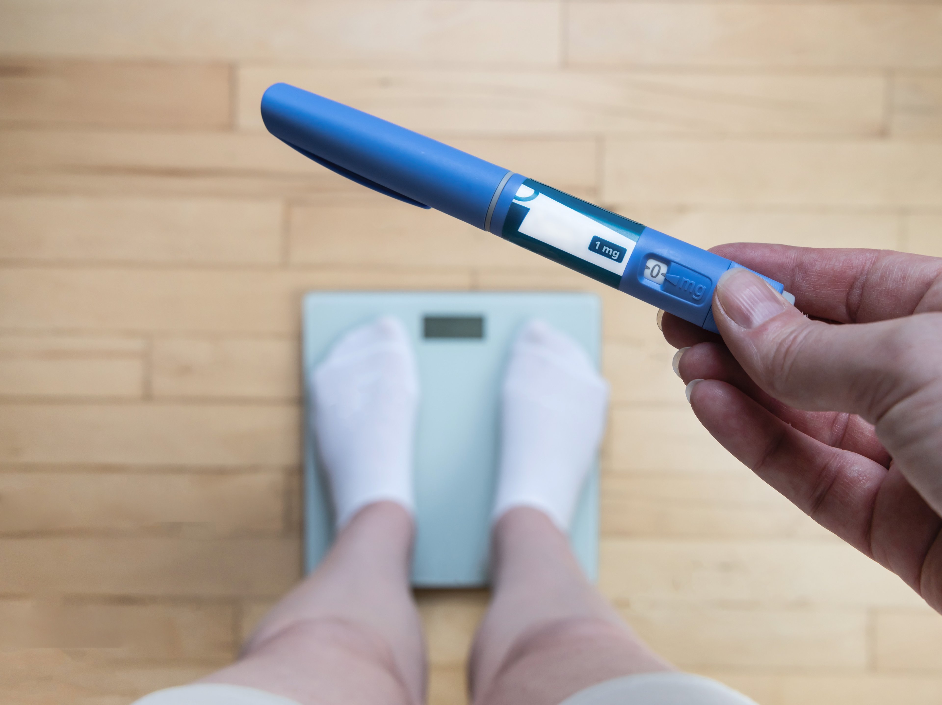 A person holds a weight-loss injector pen while standing on a scale.