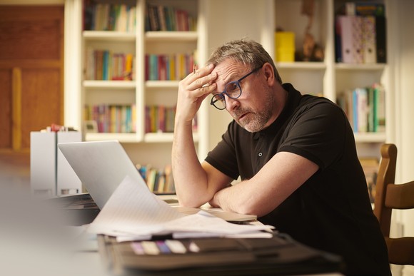 Worried Apple investor sits at a desk looking at a laptop screen with bookshelves in the background.