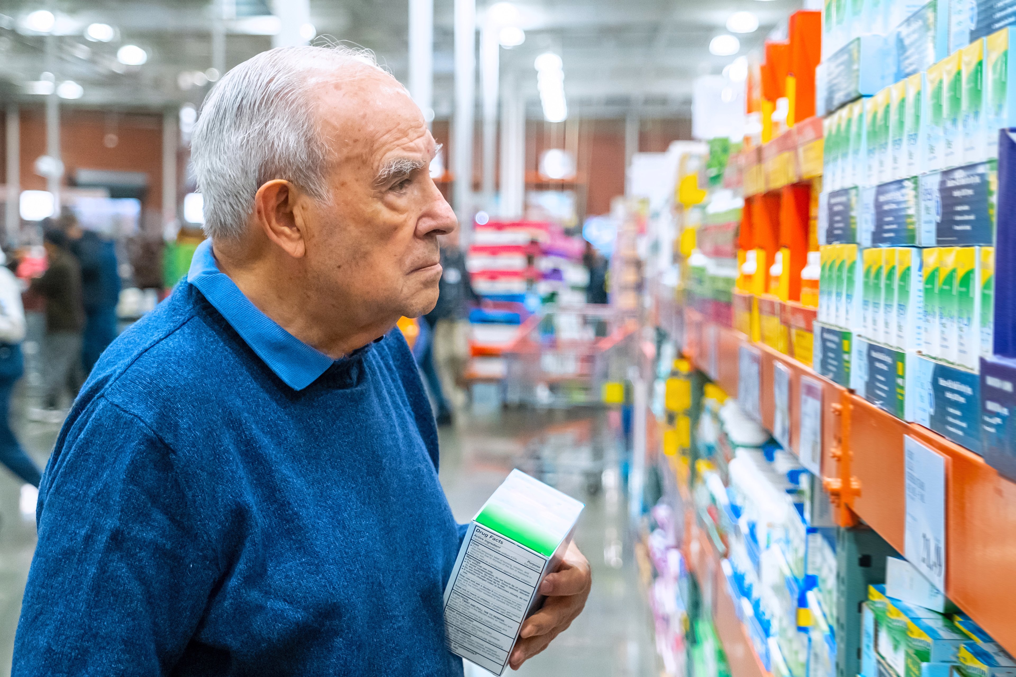 Older person dressed in collared blue shirt, studying options available on a pharmacy shelf. 