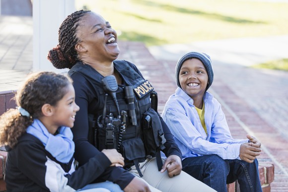 A police officer laughs while sitting and talking to two young children.