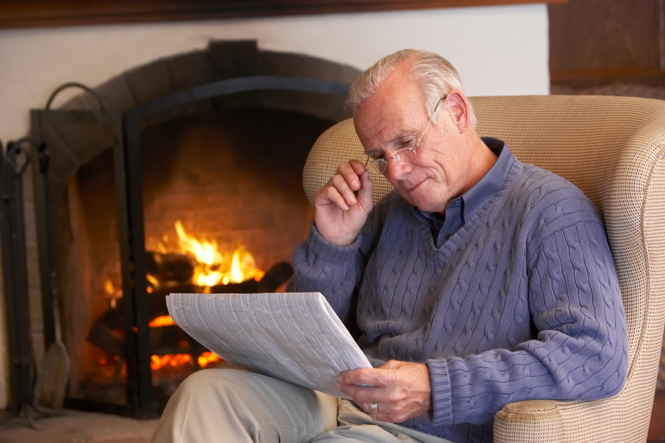 Person in lavender sweater, reading a newspaper by a fireplace. 