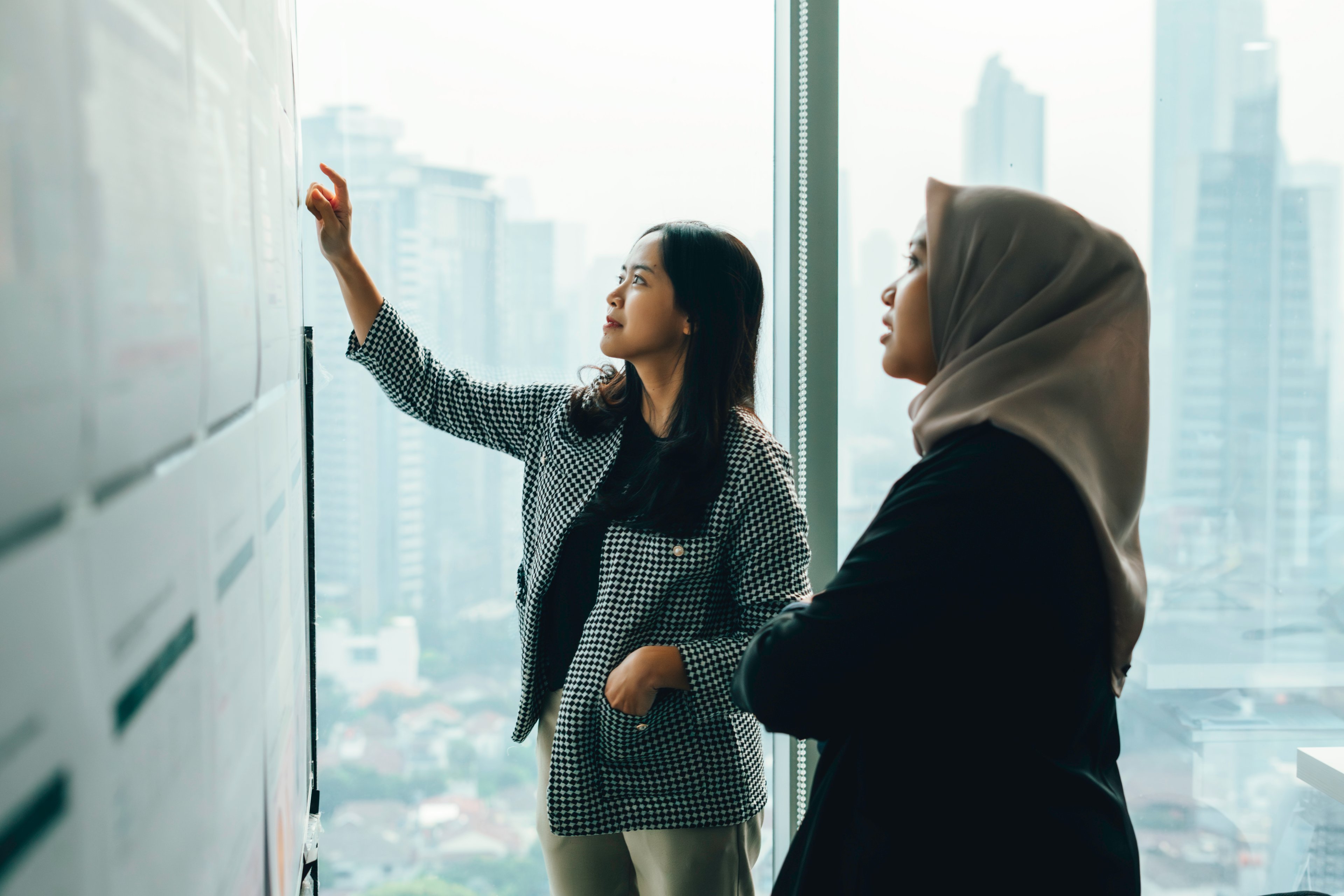 Two coworkers examine a board with postings inside their office overlooking the city.