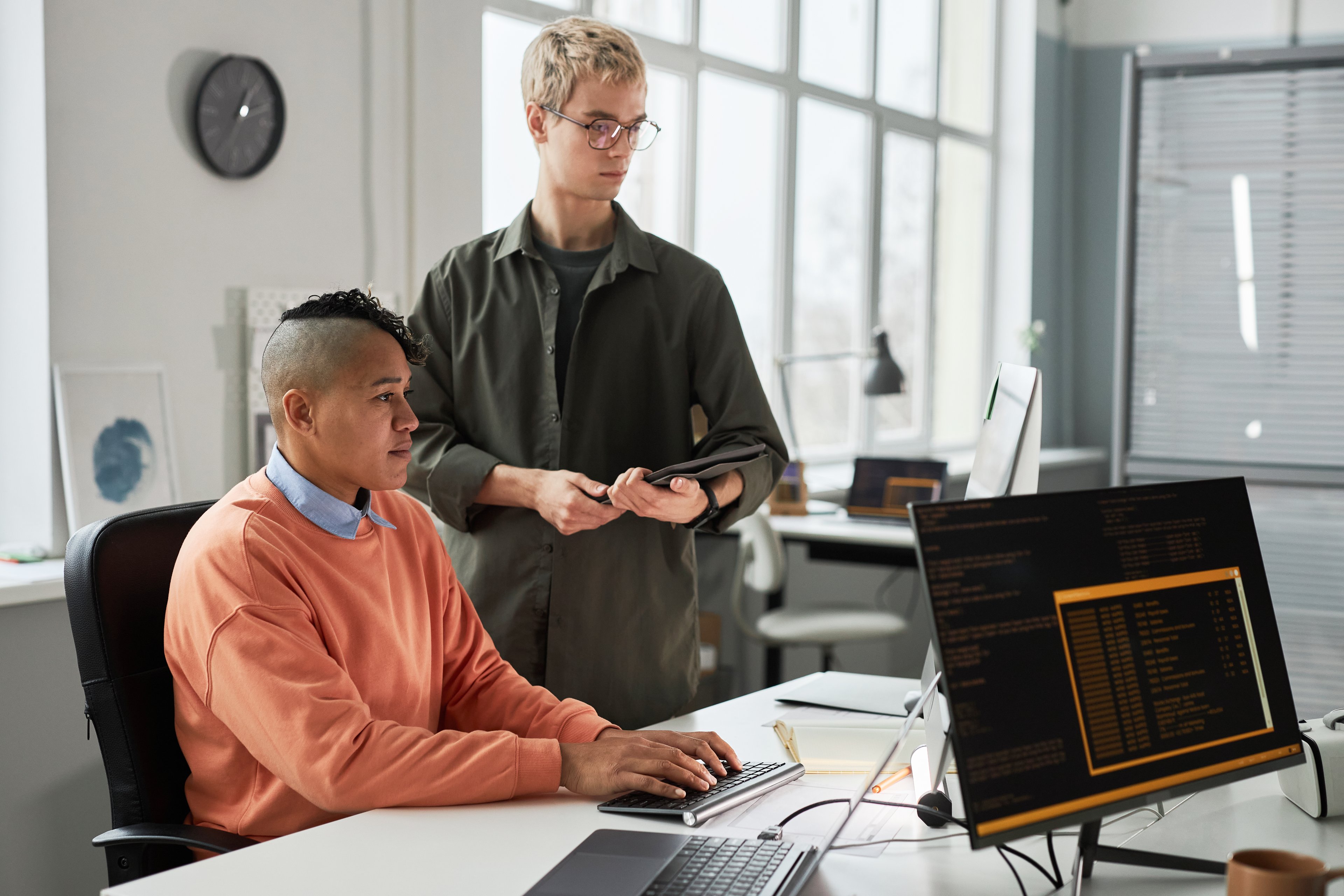 Two people (one standing, the other sitting) looking at a computer in an office setting. 