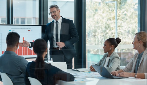 A corporate team meets in a conference room where one member appears to be referencing the stock chart behind them. 