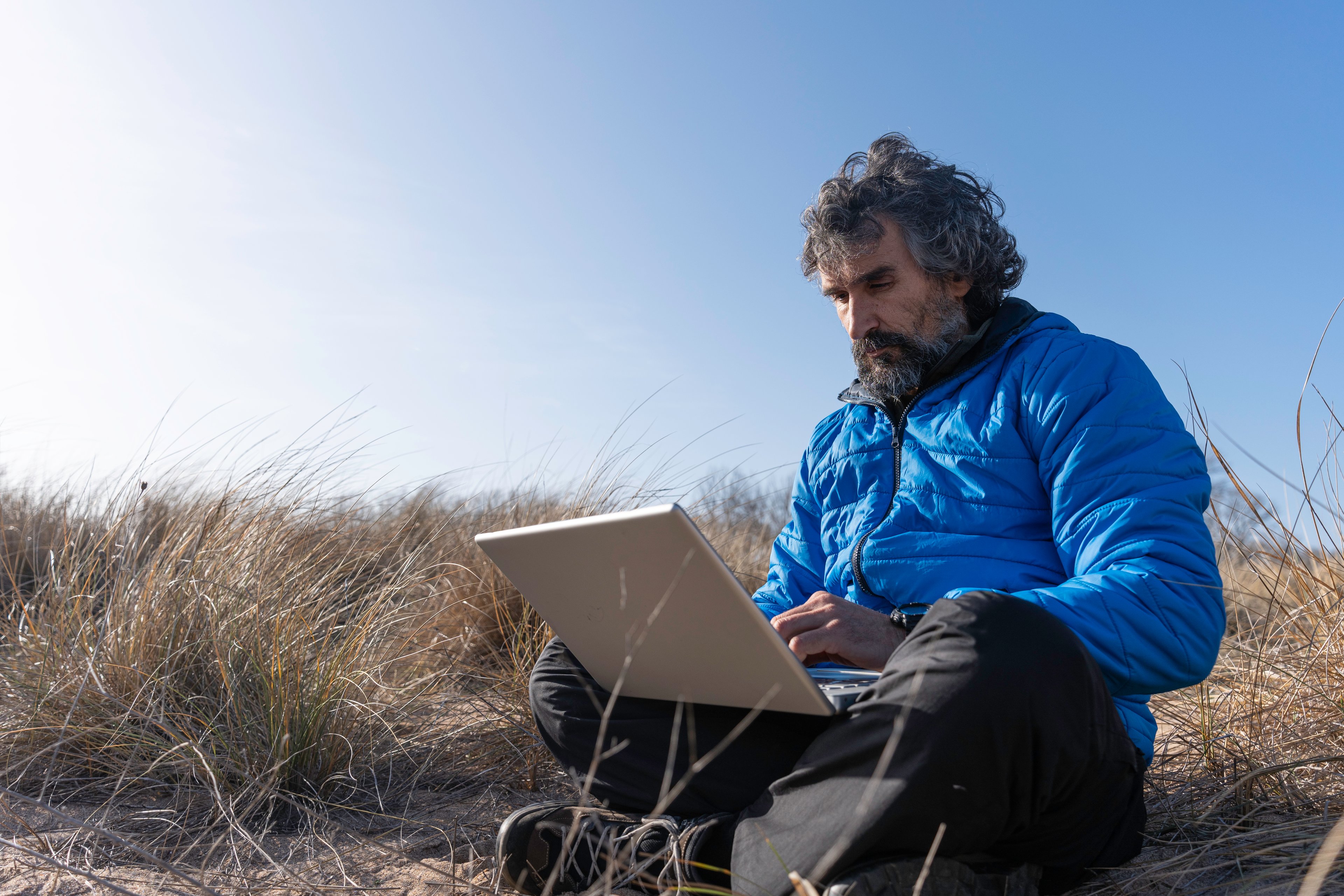 A person sits on a sandy hill and uses a laptop.