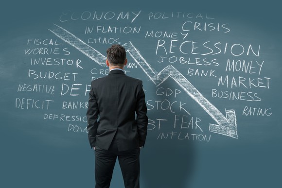 Businessperson in a suit facing a chalkboard, with words like economy, crisis, inflation, and recession written in chalk and an arrow facing downward. 