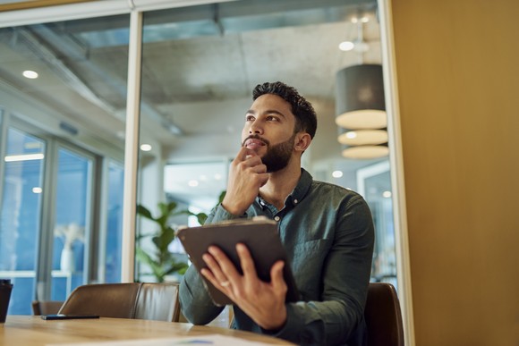 An analyst sits at a modern office table holding a tablet, appearing to be in deep thought.