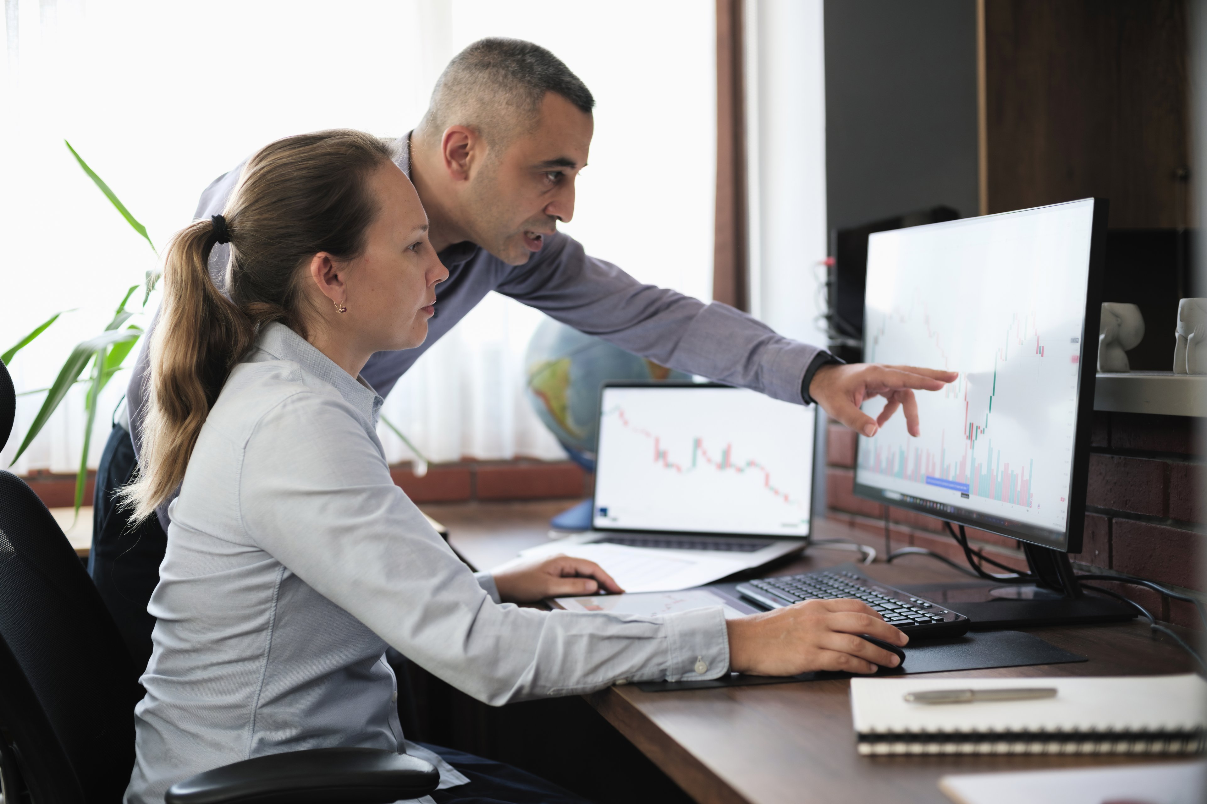 Two investors look at a pair of stock charts displayed on screens in their office as one gestures to a specific point on one of the charts.