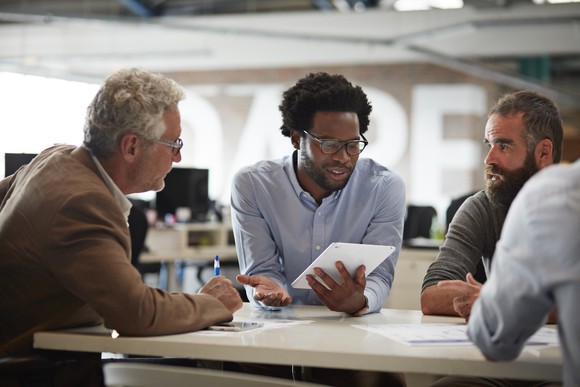A group of professionals is engaged in a focused discussion around a tablet during a business meeting.