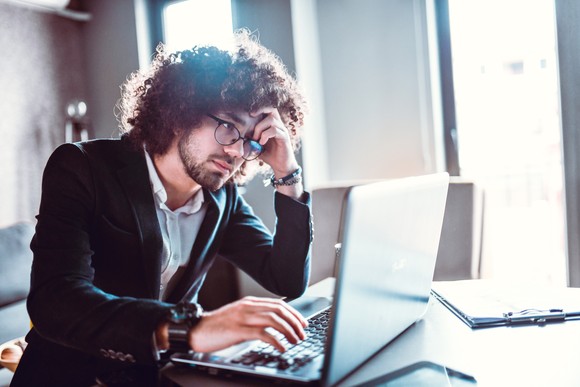 An investor holds his head in consternation as he looks at a laptop at his desk.