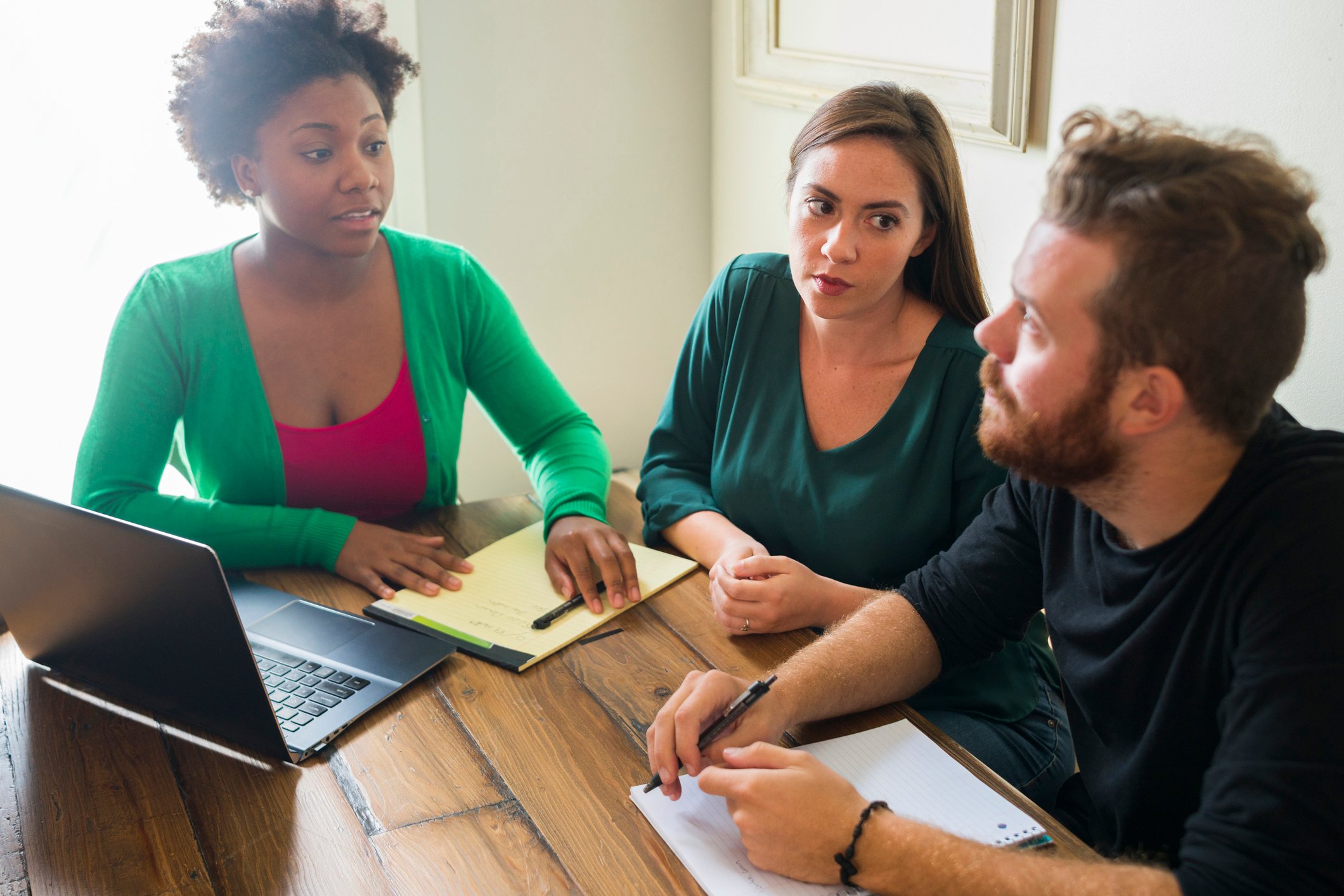 Three people seated at a table, with a laptop, while involved in a discussion.