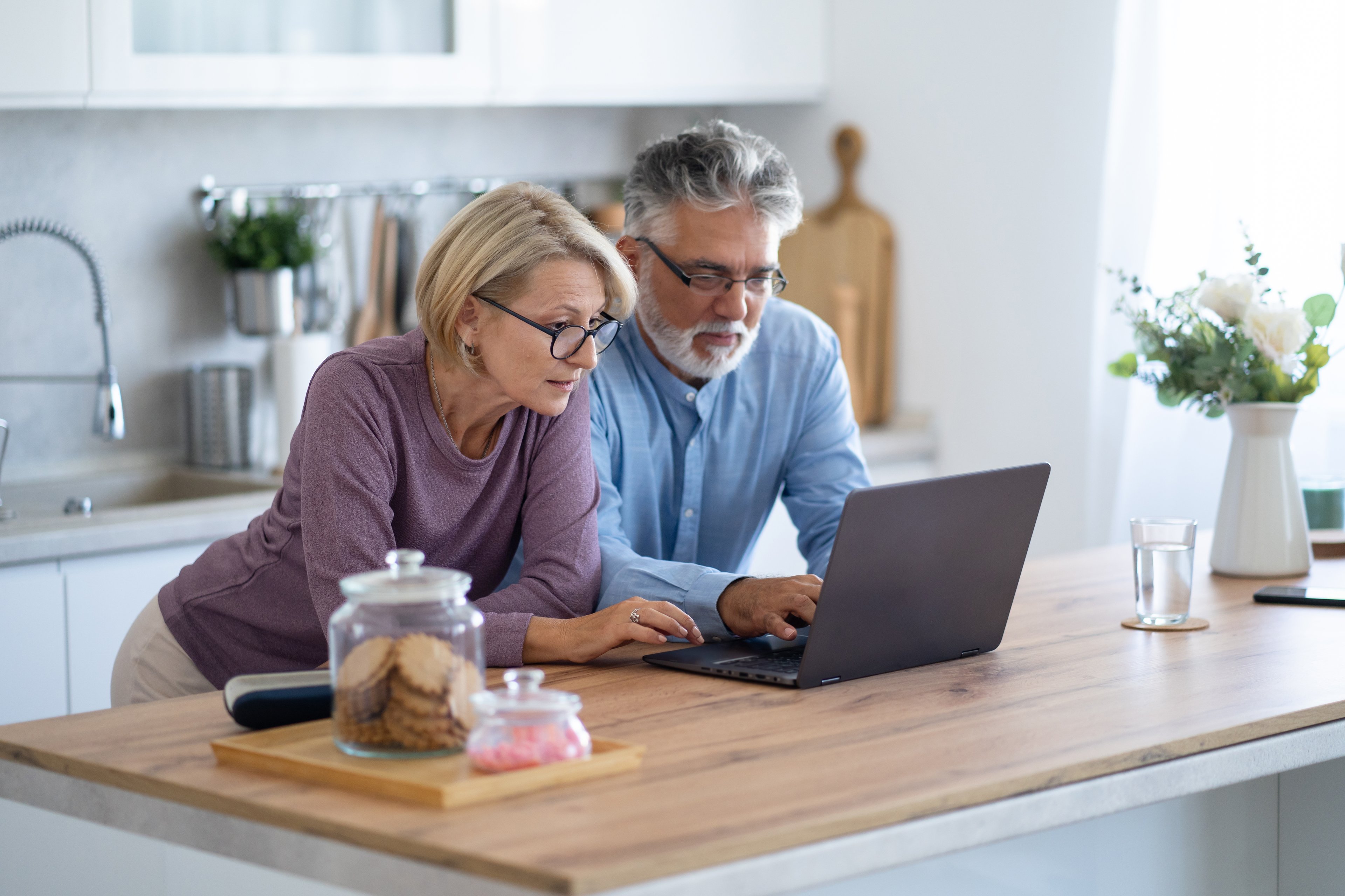 Older couple stand over their kitchen island, looking at a laptop together. 