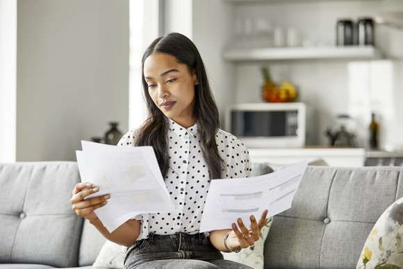An investor sits on a couch as she examines two pieces of paper with stock charts printed onto each.