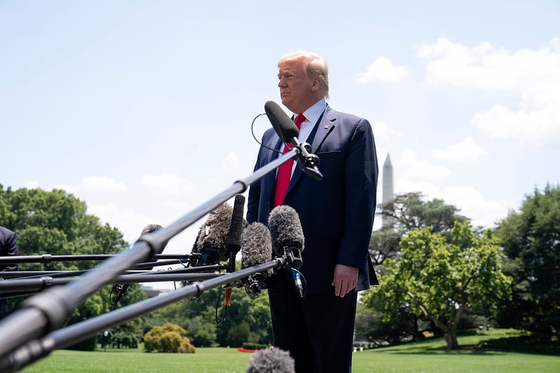 President Donald Trump standing before microphones in an outdoor setting with lots of greenery in the background.