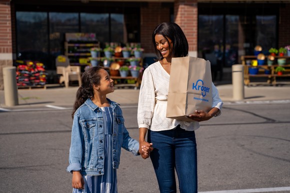 A parent and child hold hands and smile as they leave a Kroger grocery store. 