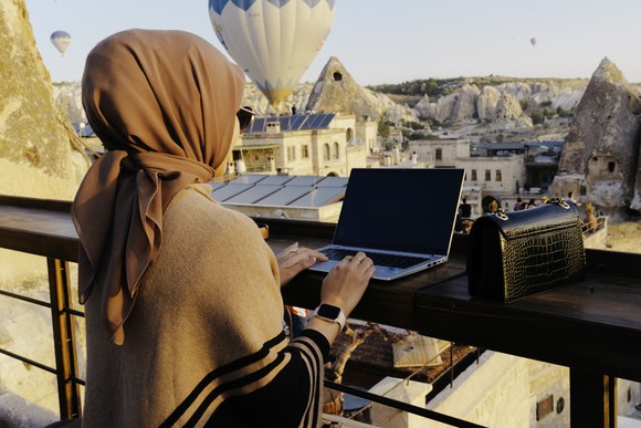 Woman in headscarf on porch on laptop with handbag. 