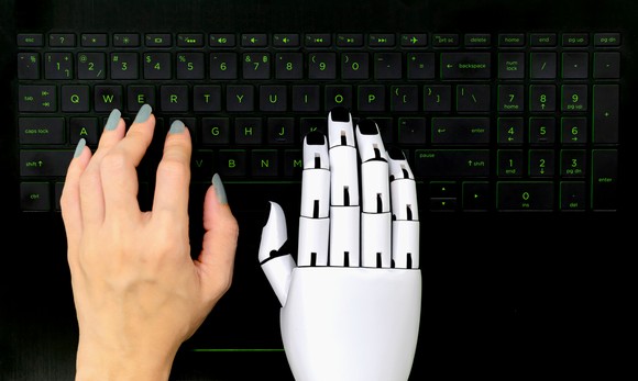 One human hand and one robotic hand use a computer keyboard.