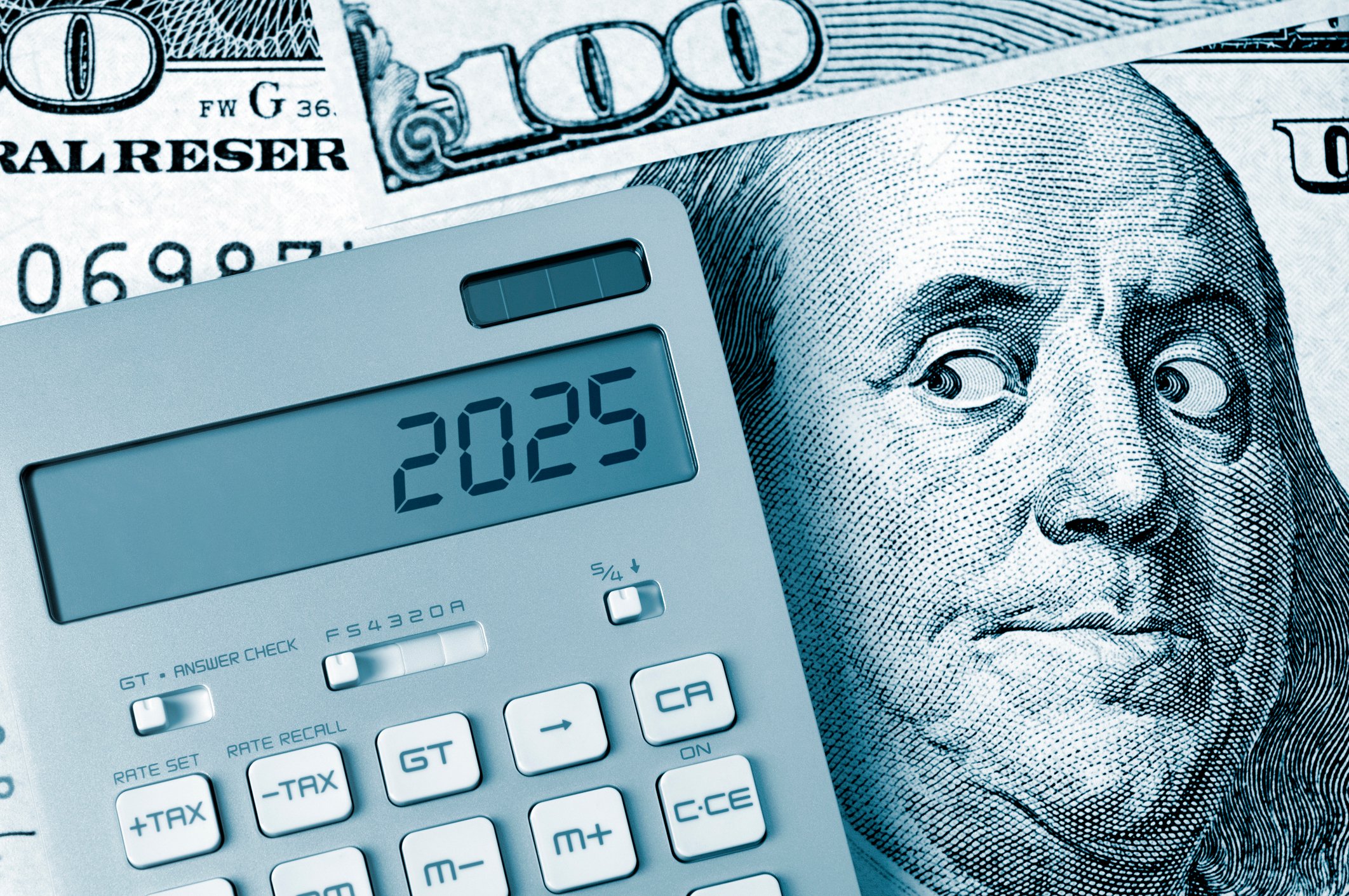 A calculator showing the number 2025 rests on a heap of hundred-dollar bills. The Ben Franklin image on one bill glances at the calculator.