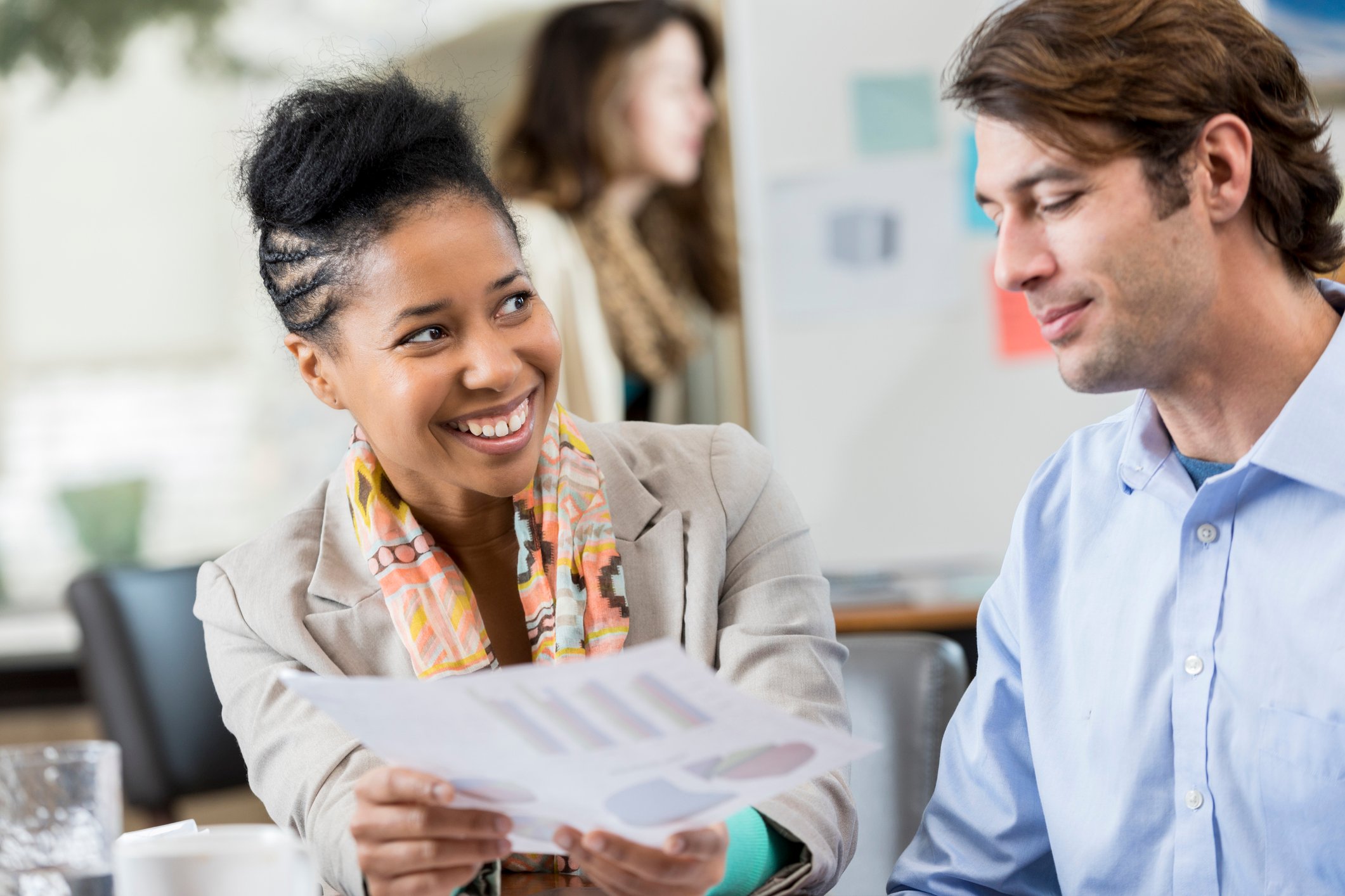 One investor smiles as she hands a paper with data to another investor as they sit in an office meeting room.
